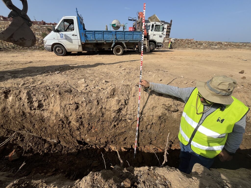 Arqueólogo midiendo con mira la profundidad de un sondeo en una obra, con maquinaria y edificios al fondo